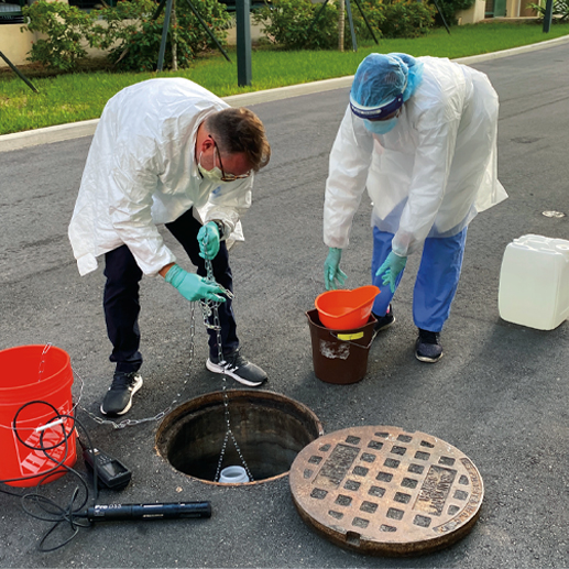 Two people wearing protective scrubs, masks, and gloves collect wastewater sample from manhole on a road, draining a residential building.  A collecting bucket and container lay on the road near them.